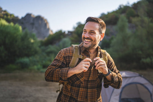 Man with a backpack standing outdoors near a tent with greenery in the background