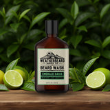 Bottle of Weatherbeard Supply Co's Emerald Oasis Beard Wash Sitting On A Wooden Table Surrounded By Greenery And Lime Slices