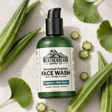 Weatherbeard Supply Co. Botanical Peptide Face Wash in a green bottle with a black pump, surrounded by fresh green okra and ginkgo leaves on a light stone background.