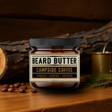 Jar of beard butter labeled 'Campside Coffee' on a wooden surface with coffee beans and a log in the background.