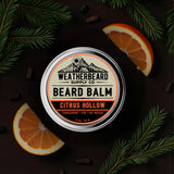 Beard balm container with 'Weatherbeard Supply Co' branding, surrounded by tangerine slices and green leaves on a dark background.