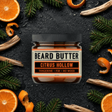 Jar of  'Citrus Hollow' Beard Butter surrounded by tangerine slices, fir needle, and wood chips on a dark background