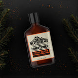Bottle of Weatherbeard Supply Co. Citrus Hollow Conditioner on a dark background with fir needle branches.