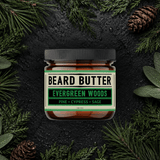 Jar of beard butter labeled 'Evergreen Woods' surrounded by pine branches and cones on a dark background