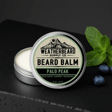 Palo Peak Beard Balm on a dark background with spearmint mint leaves and juniper berries surrounding it.