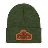 An olive green ribbed knit beanie hat with a brown leather patch on the front cuff that reads 'WEATHERBEARD SUPPLY CO' with a mountain graphic.