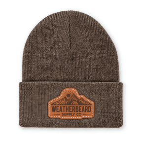 A brown ribbed knit beanie hat with a folded cuff, featuring a brown leather patch on the front that reads 'WEATHERBEARD SUPPLY CO' with a mountain graphic, shown on a plain white background.