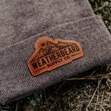 A close-up of a dark heather gray knit beanie cuff featuring a brown leather patch with a mountain graphic and the text 'WEATHERBEARD SUPPLY CO'. The beanie is placed outdoors on a bed of dry pine needles and soil.