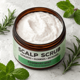 An open amber jar of scalp scrub with white cream inside, surrounded by mint leaves and rosemary sprigs on a light stone surface. The label reads 'SCALP SCRUB' and 'ROSEMARY MINT With Aloe & Fenugreek'.